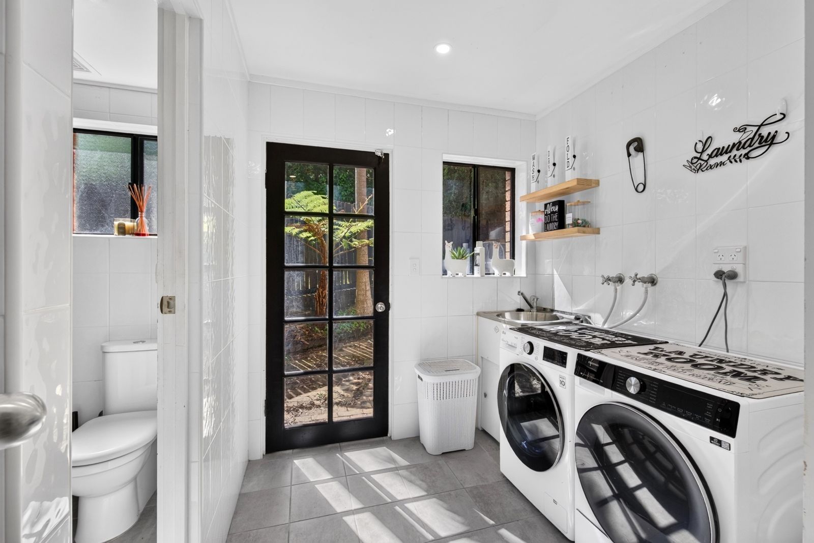 Bright white laundry with front-loading washer and dryer, timber shelves and a black glass door to the garden